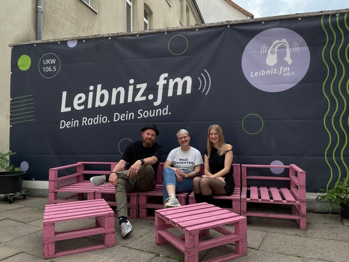 Vor einem Leibniz.fm Banner sitzen drei Leute auf pinken Paletten und lächeln in die Kamera.