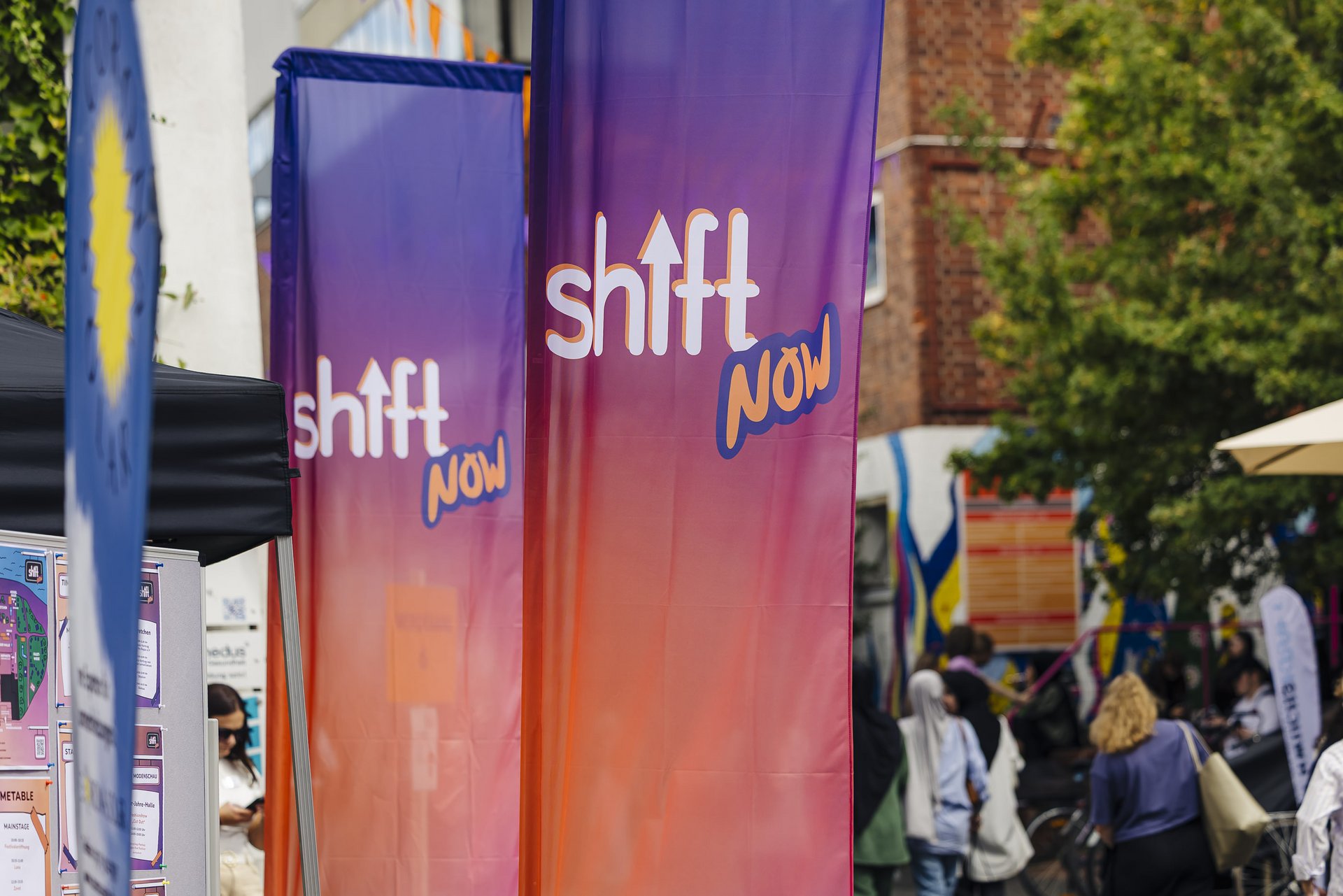 Man sieht zwei SHIFT NOW Flaggen in lila,oangenem Farbverlauf auf einem Festivalgelände.