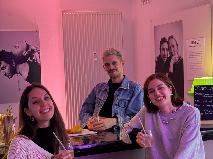 Man sieht drei Personen an einer Bar. Vor ihr sitzen zwei Frauen mit einem Getränk in der Hand. Mittig hinter der Bar steht ein Mann. Alle lachen in die Kamera.