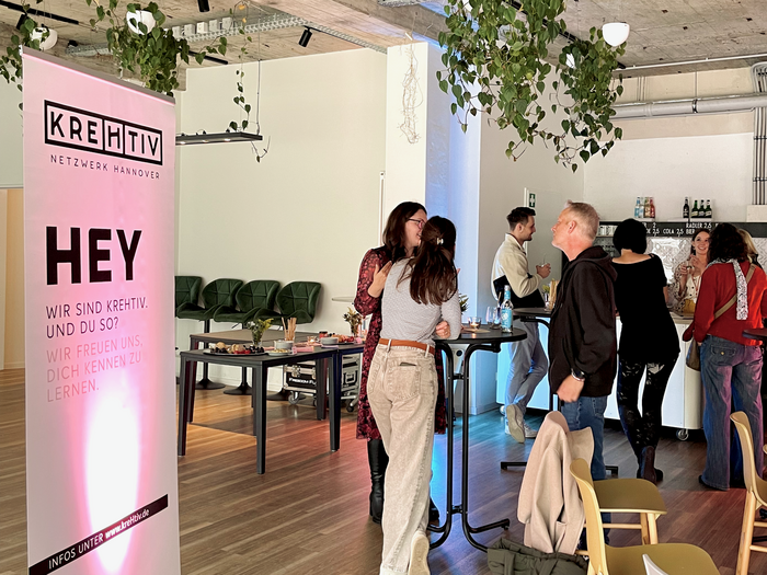 Menschen stehen in einem Foyer an Stehtischen und unterhalten sich. Man sieht links im Bild einen Banner vom kreHtiv Netzwerk.