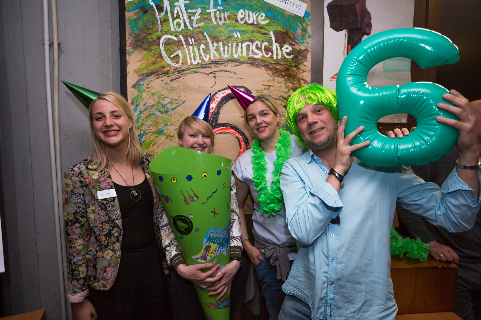 4. Menschen stehen vor einer Fotowand. Eine hat eine Schultüte in der Hand, ein anderer hat eine grüne Perücke auf und hält eine 6 hoch.