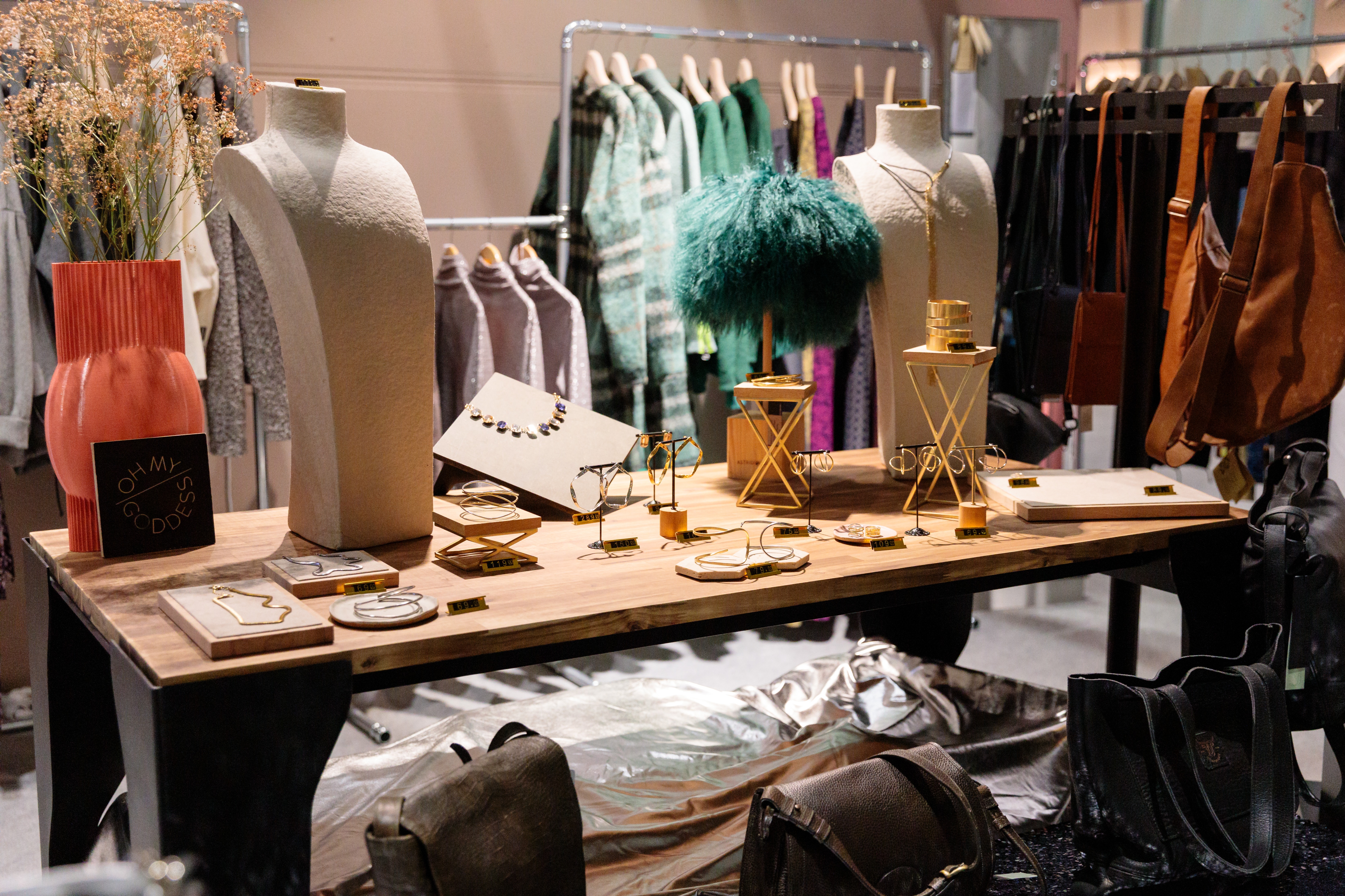 Auf einem Tisch liegen verschiedene Mode-Accessoires, darunter Halsketten. Vor dem Tisch stehen Handtaschen, hinter dem Tisch hängt Kleidung. 
